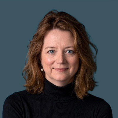 A woman with light skin and shoulder-length wavy brown hair wearing a black turtleneck sweater stands in front of a plain dark blue background, smiling softly at the camera.